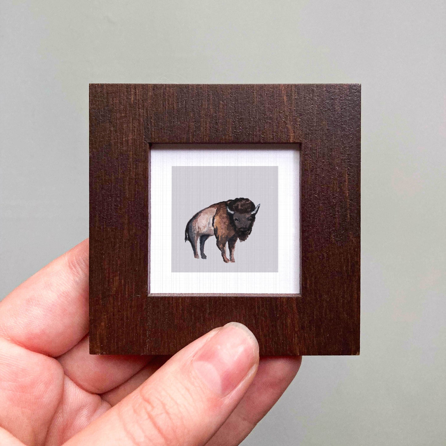 A hand is holding a small wooden frame containing a picture of a bison.
