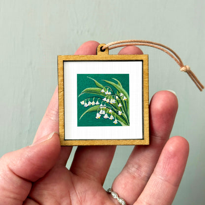 A hand holding a small wooden frame containing a picture of lily of the valley flowers.