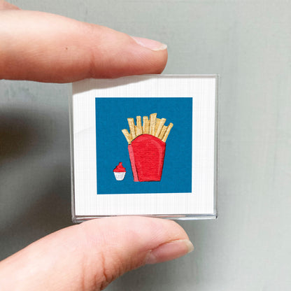 A person's hand holding a small square with a red container and french fries depicted on it.
