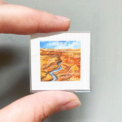 A person's hand holding a small square picture of a desert landscape with a winding river.