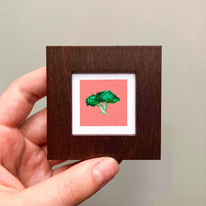 A hand is holding a small square frame containing a vibrant image of a green broccoli floret against a red background.