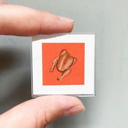 A hand is holding a small square with an illustration of a roasted chicken on it.