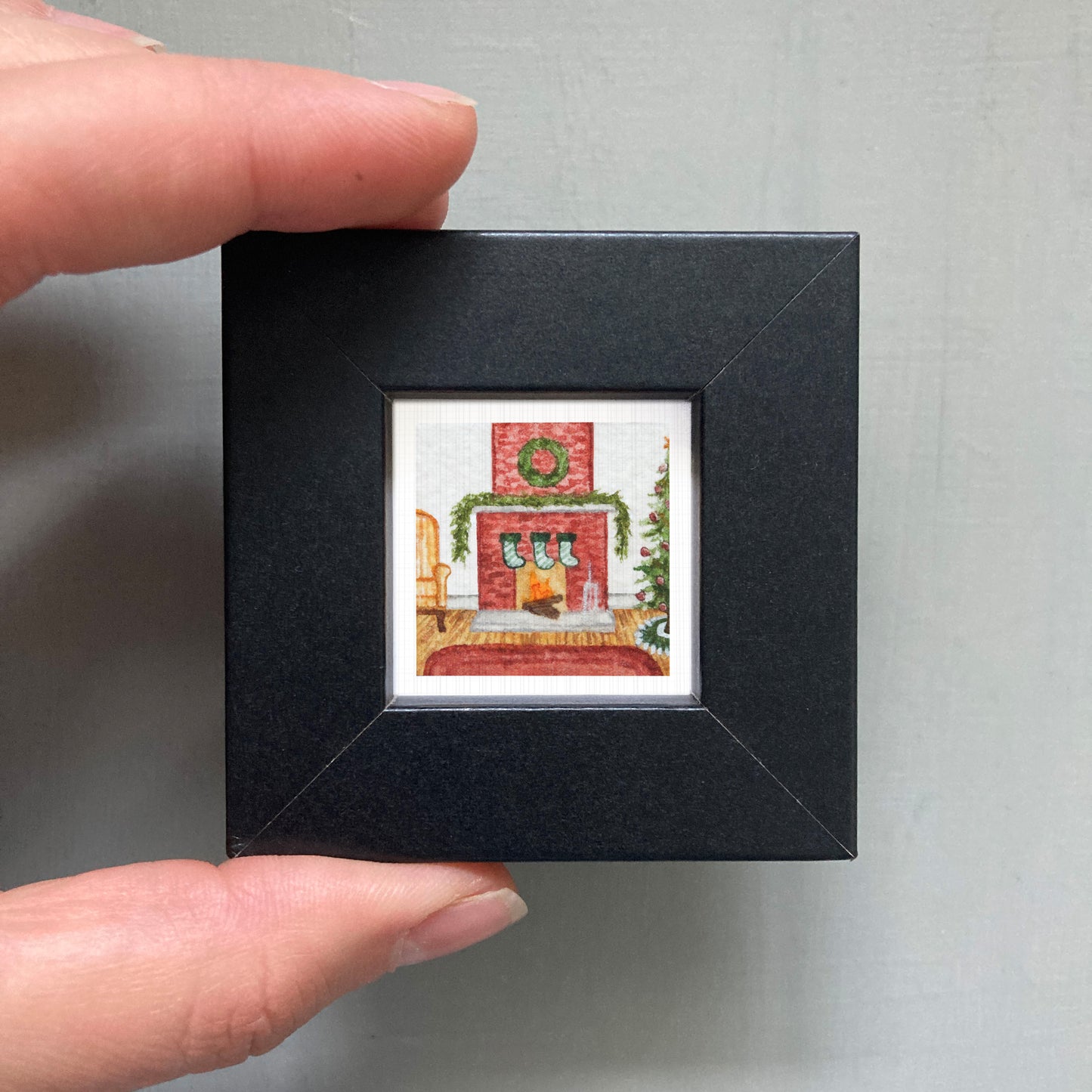 A hand is holding a small black picture frame containing a small, colorful painting of a fireplace with a wreath on the mantel.