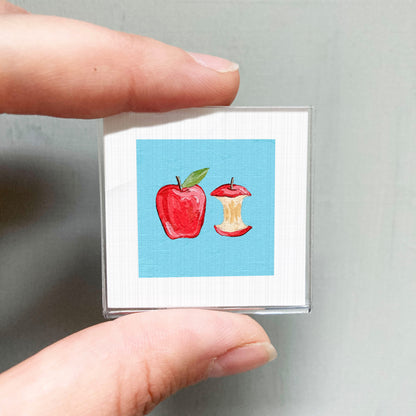 A person's hand holding a small square with a painting of an apple and an apple core on it.