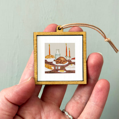 A person's hand holding a small wooden frame containing a framed illustration of a table with various food items, such as a turkey, bread, and other dishes.