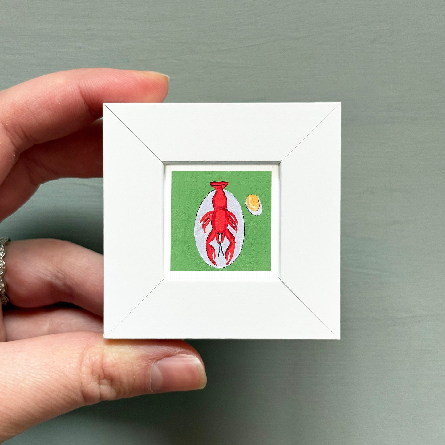 A hand is holding a small white picture frame containing an illustration of a red lobster.