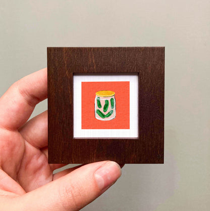 A hand is holding a small wooden frame containing a framed illustration of a jar of pickles.