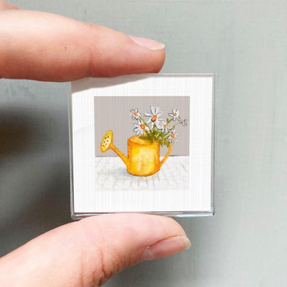 A person's hand holding a small square picture of a yellow watering can with white flowers.