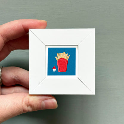 A hand is holding a small white frame containing a framed image of a red cup with french fries and a cupcake.