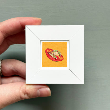 A hand is holding a small white square frame containing a vibrant illustration of a red basket filled with food.