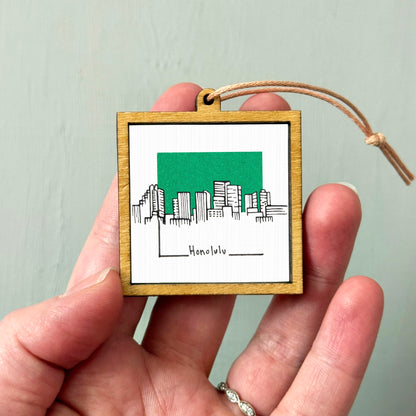 A hand holding a small wooden frame with a picture of the skyline of Honolulu, Hawaii.