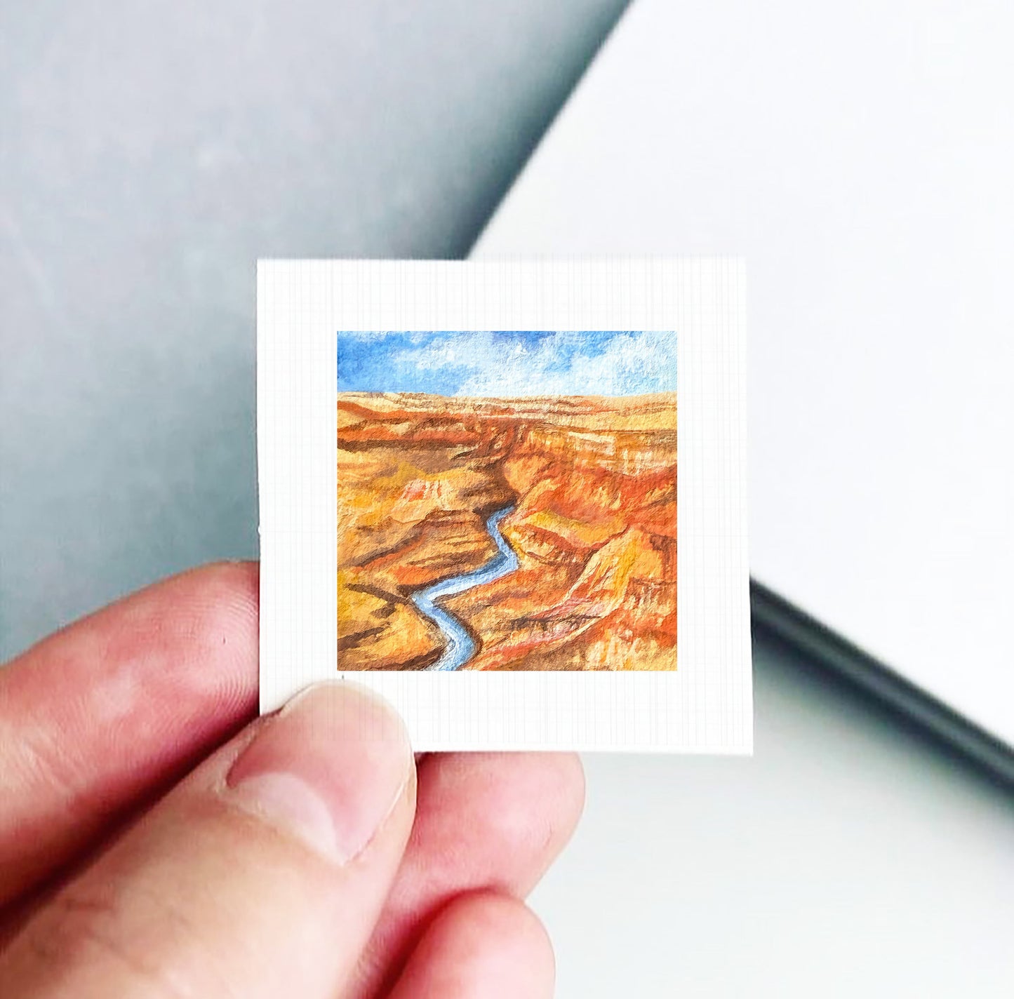 A hand is holding a small square piece of paper with a painting of a desert landscape featuring a winding river.