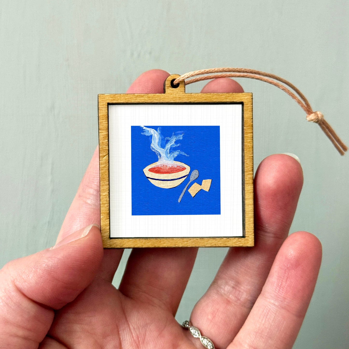 A person's hand holding a small wooden frame containing an illustration of a bowl of soup with steam rising from it.