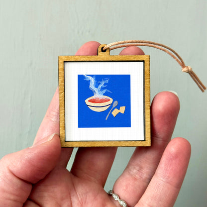 A person's hand holding a small wooden frame containing an illustration of a bowl of soup with steam rising from it.