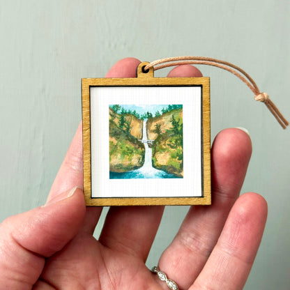 A hand holding a small wooden frame containing a framed watercolor painting of a waterfall.