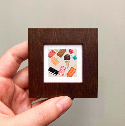 A hand is holding a small square picture frame containing a colorful illustration of various ice cream flavors.