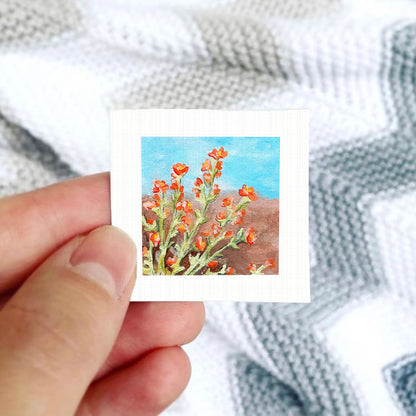 A hand is holding a small square picture of a field of red flowers against a blue sky background.