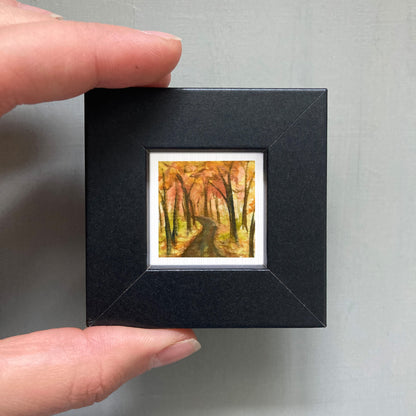 A person's hand holding a small, square-shaped black frame containing a miniature painting of a forest path with autumn foliage.