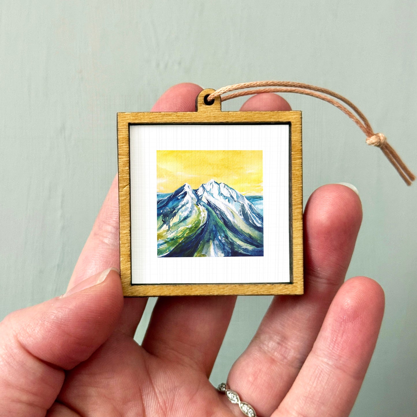 A hand holding a small wooden frame containing a painting of a mountain landscape.