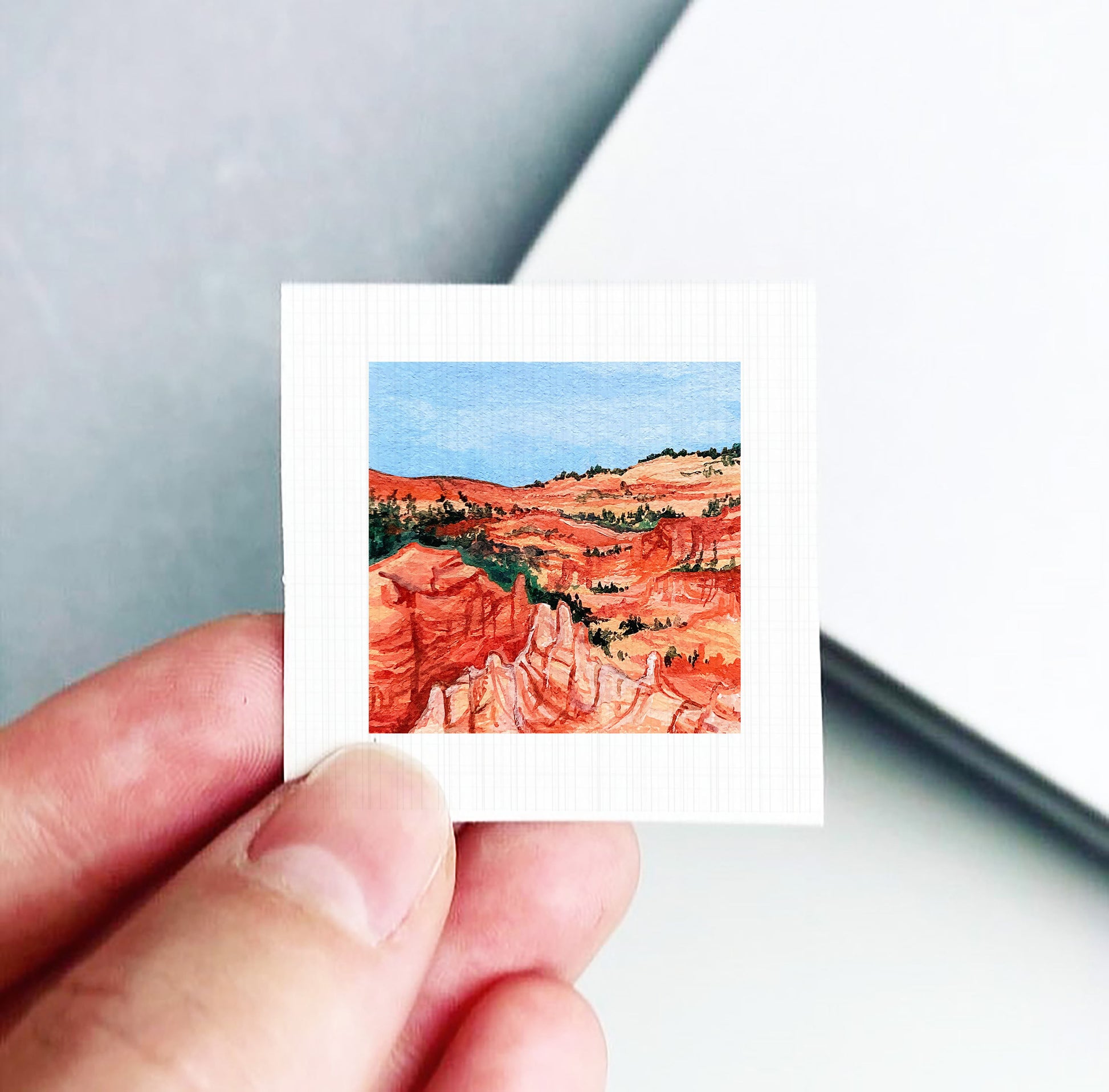 A hand holding a small square-shaped piece of paper with a painting of a desert landscape on it.