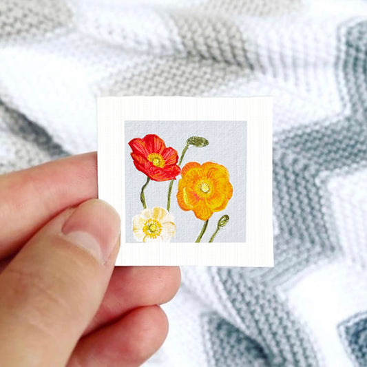 A hand is holding a small square card with a painting of two flowers, one red and one yellow, against a background of a white and gray striped fabric.