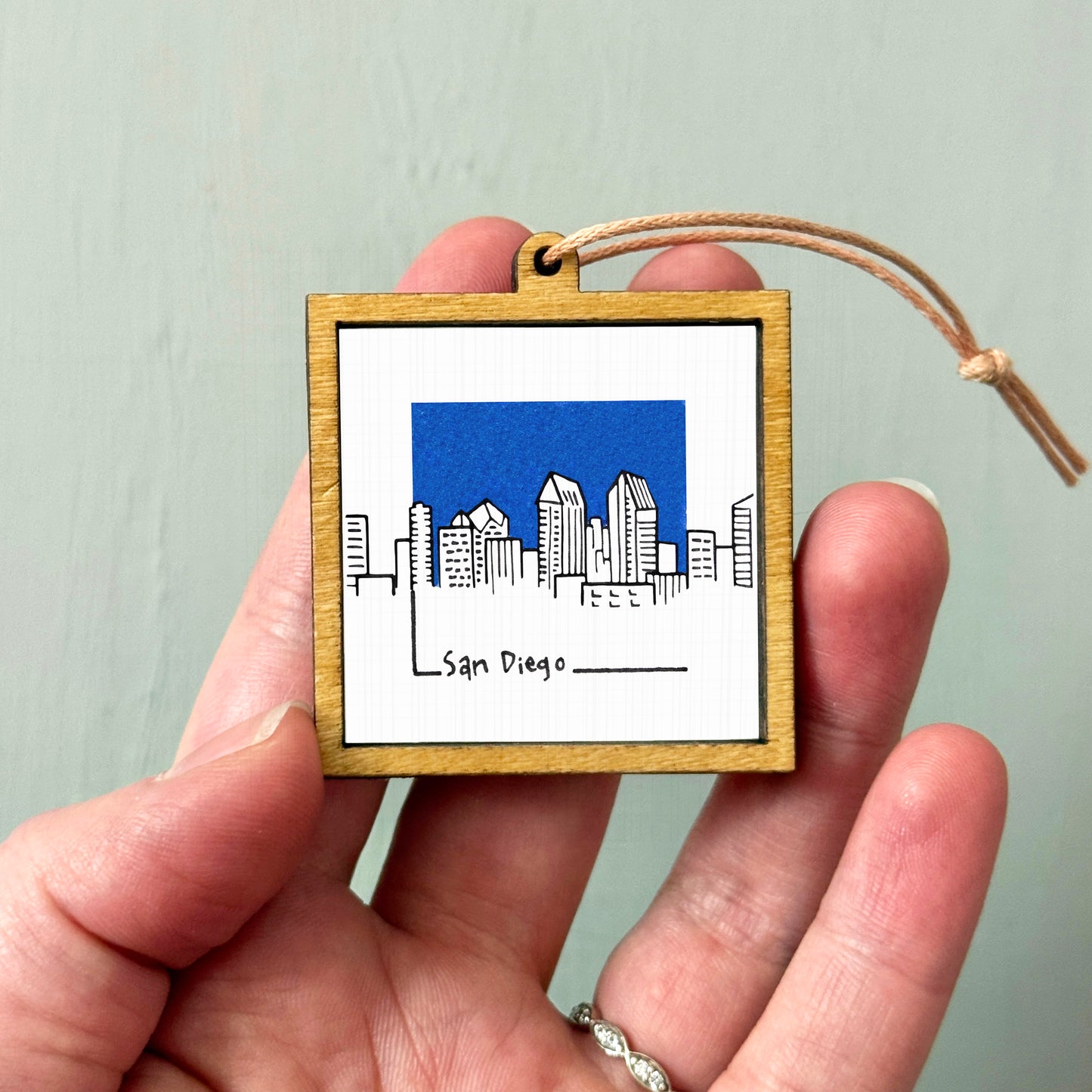 A hand holding a small wooden frame with a picture of a city skyline and the text "San Diego" written on it.