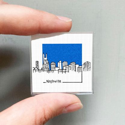 A person's hand holding a small square magnet with a drawing of a city skyline and the word "Nashville" written below it.