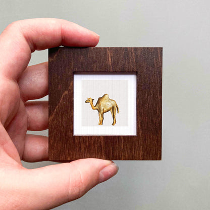 A hand is holding a small wooden frame containing a picture of a camel.