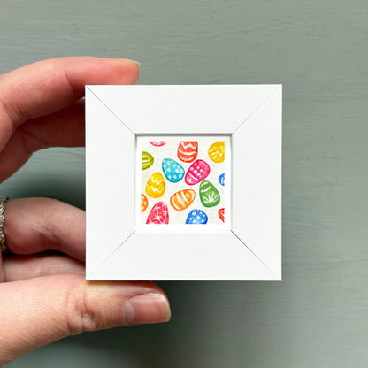A hand is holding a small white square frame containing a colorful pattern of Easter eggs.