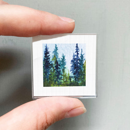 A person's hand holding a small square picture of a forest scene with tall trees and a blue sky.
