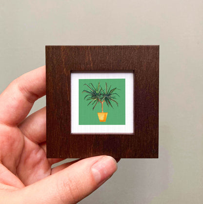 A hand is holding a small wooden frame containing a framed illustration of a potted plant with green leaves.