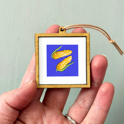 A hand holding a small wooden frame containing a framed image of corn on the cob.