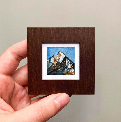 A hand is holding a small wooden frame containing a miniature painting of a mountain landscape.