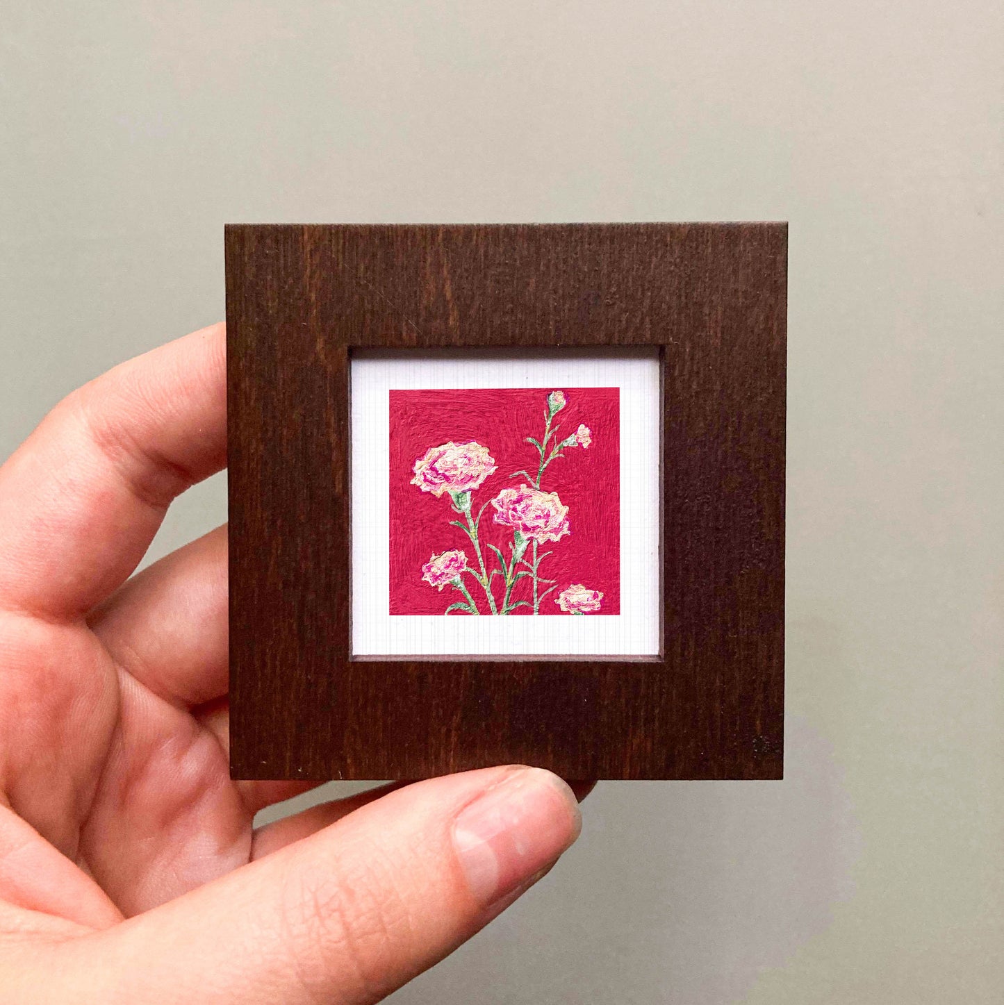 A hand is holding a small wooden frame containing a framed painting of pink flowers against a red background.