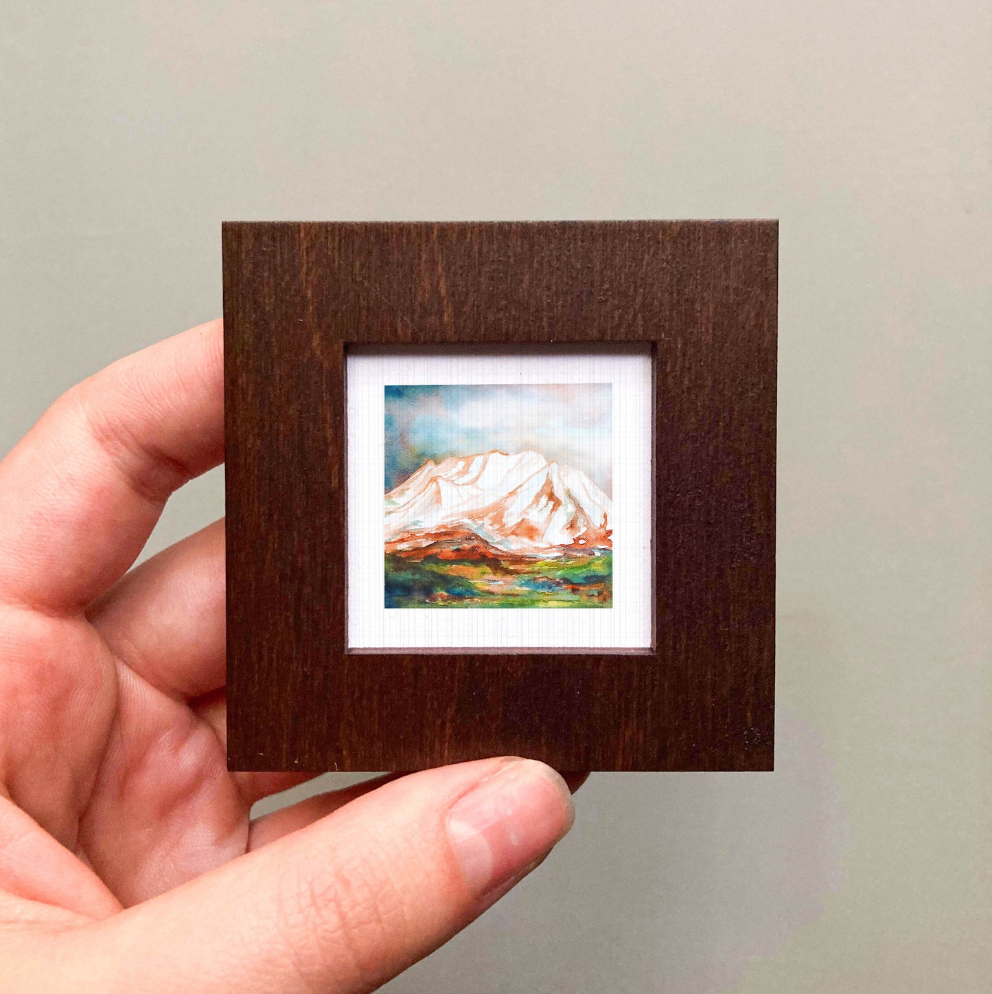 A hand is holding a small wooden frame containing a painting of a mountain landscape.