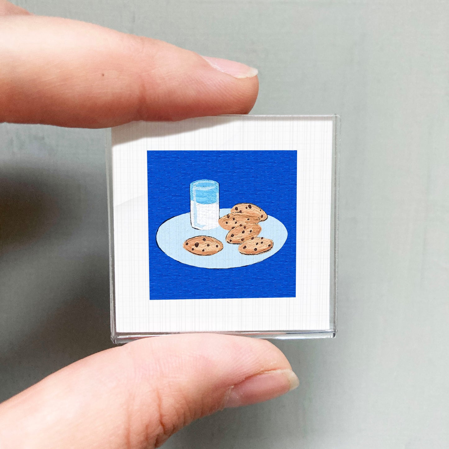 A person's hand is holding a small square with an illustration of a plate of cookies and a glass of milk on it.