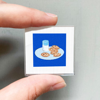 A person's hand is holding a small square with an illustration of a plate of cookies and a glass of milk on it.