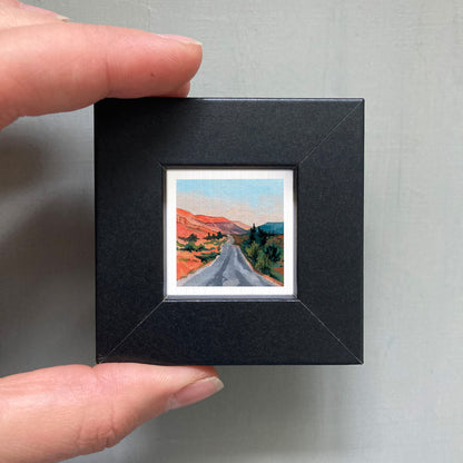 A person's hand holding a small black picture frame containing a miniature painting of a winding road through a desert landscape.