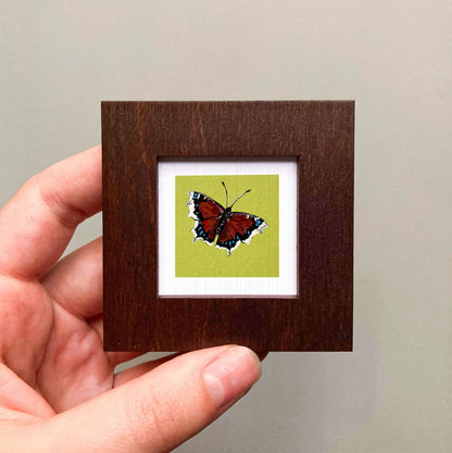 A hand is holding a small wooden frame containing a framed image of a red butterfly with black and white markings.