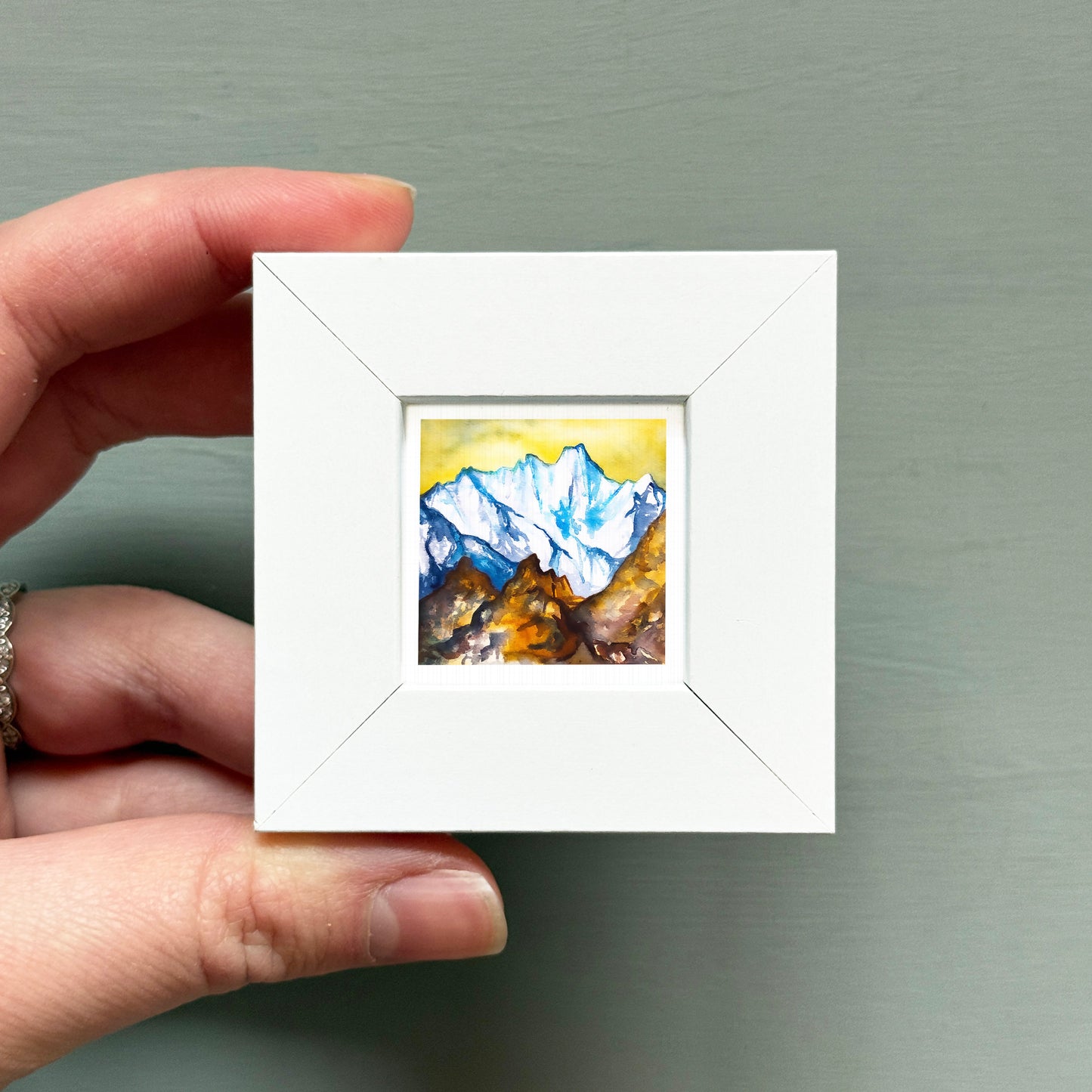 A hand is holding a small white square frame containing a watercolor painting of a mountain landscape.