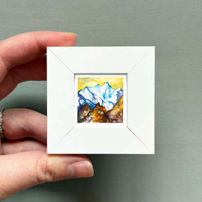 A hand is holding a small white square frame containing a watercolor painting of a mountain landscape.