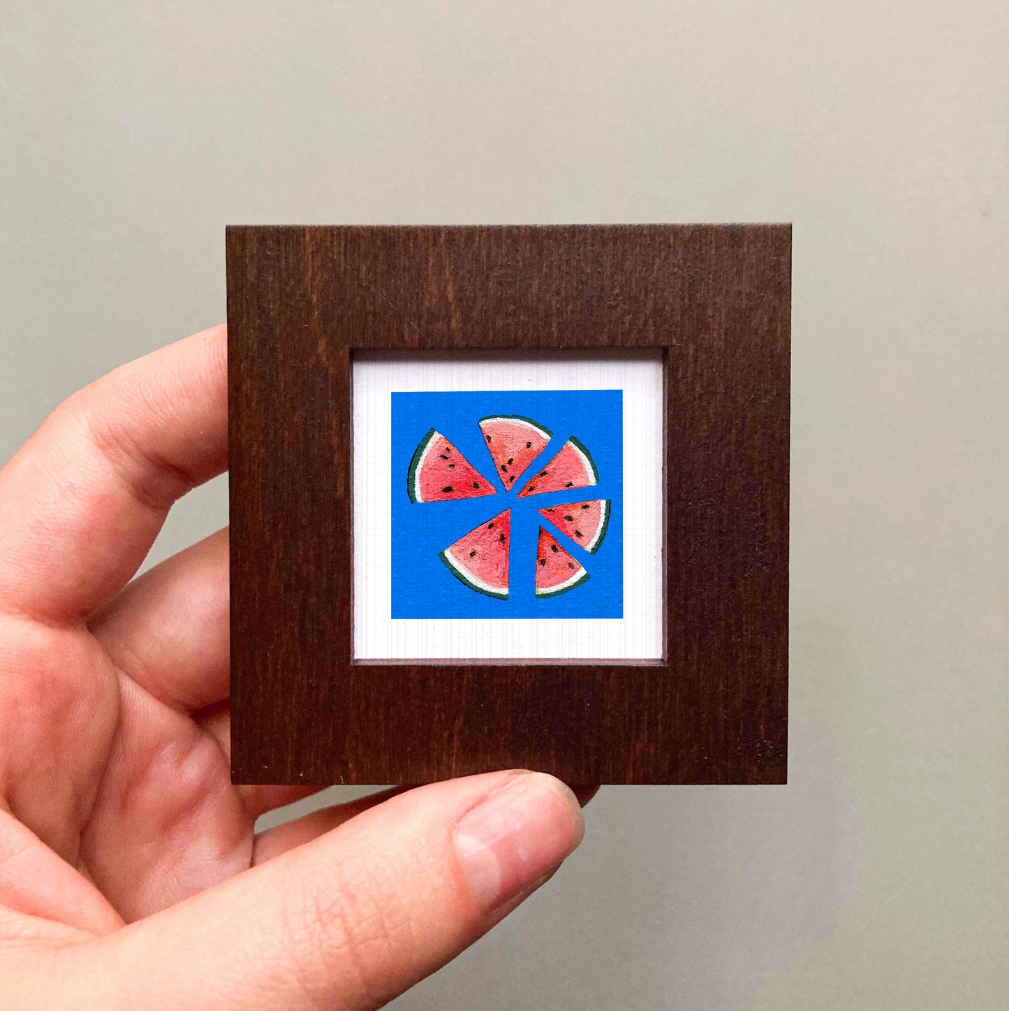 A hand is holding a small wooden frame containing a framed image of watermelon slices.