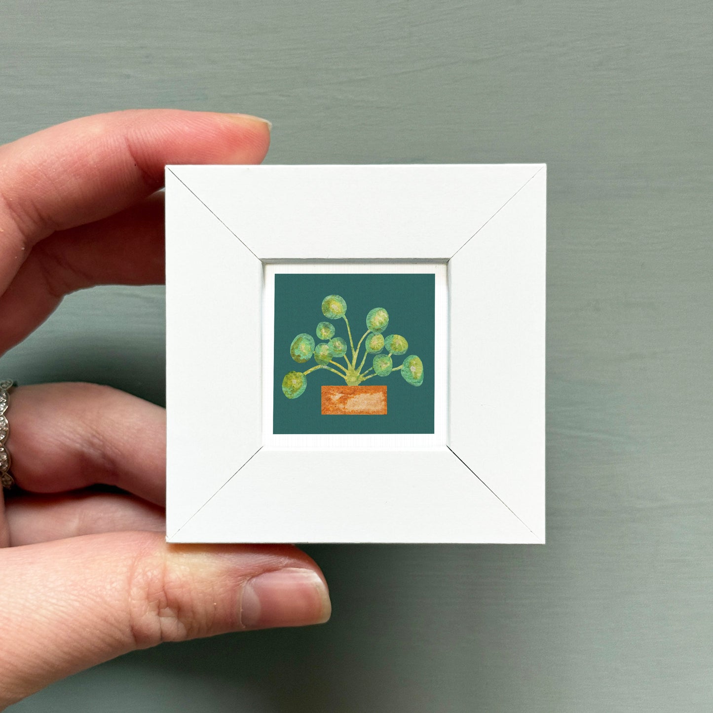 A hand is holding a small white picture frame containing a painting of a plant with green leaves and a brown pot.