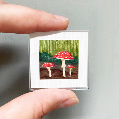 A person's hand is holding a small framed picture of three red mushrooms against a green background.