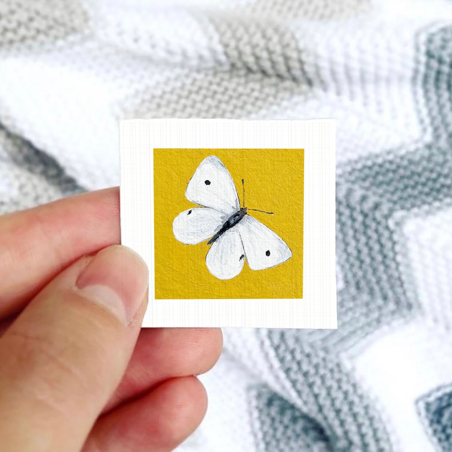 A hand is holding a small square card with a white butterfly painted on it against a background of a gray and white striped fabric.