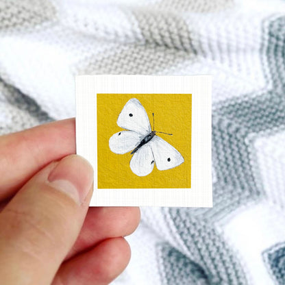 A hand is holding a small square card with a white butterfly painted on it against a background of a gray and white striped fabric.