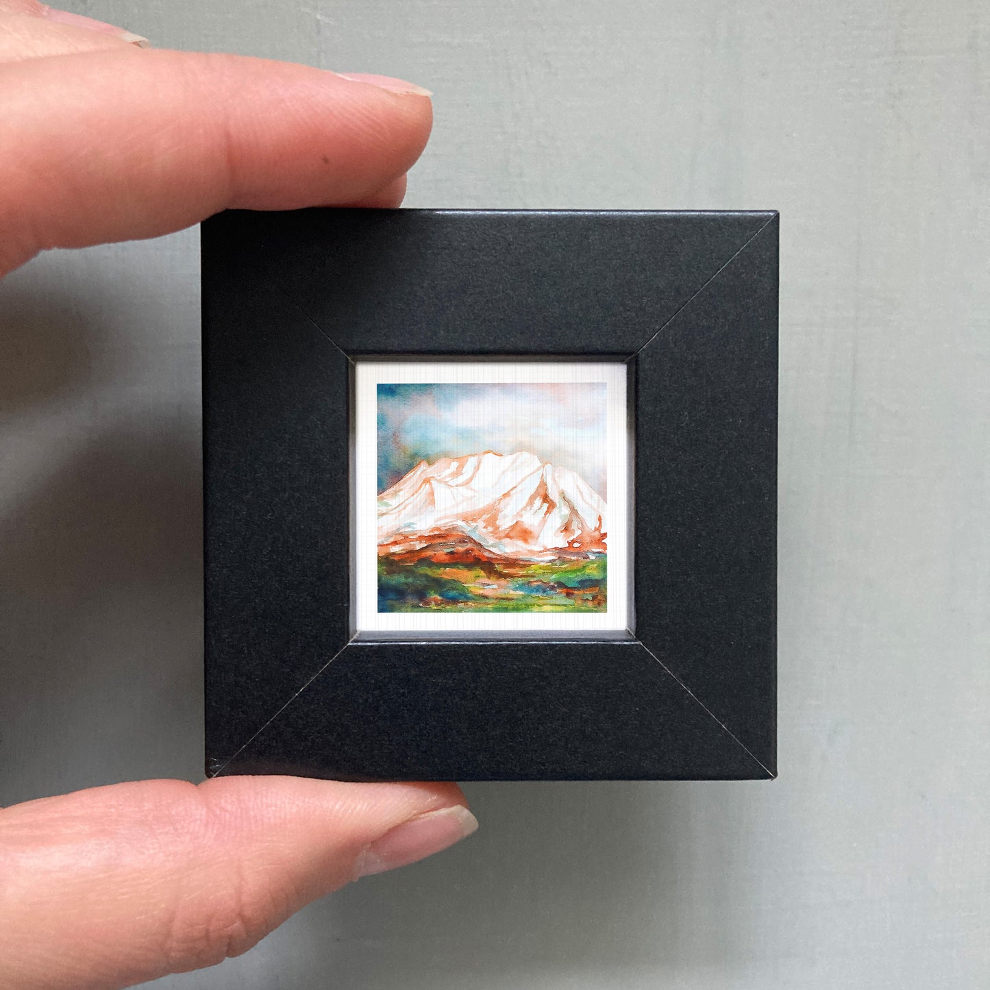 A person's hand holding a small black frame containing a framed painting of a mountain landscape.