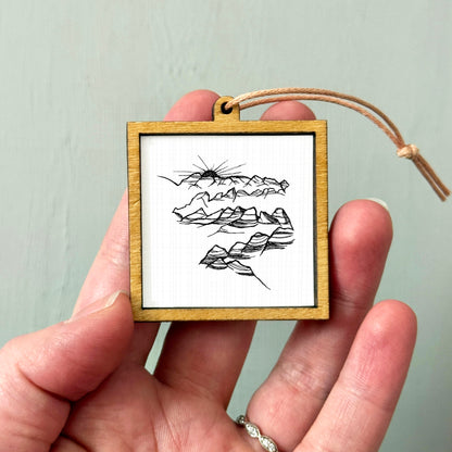 A hand holding a small wooden frame containing a black and white sketch of mountains.