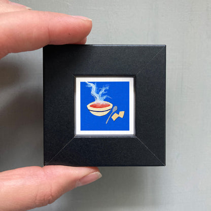 A hand is holding a small black square frame containing a vibrant illustration of a bowl of soup with steam rising from it.