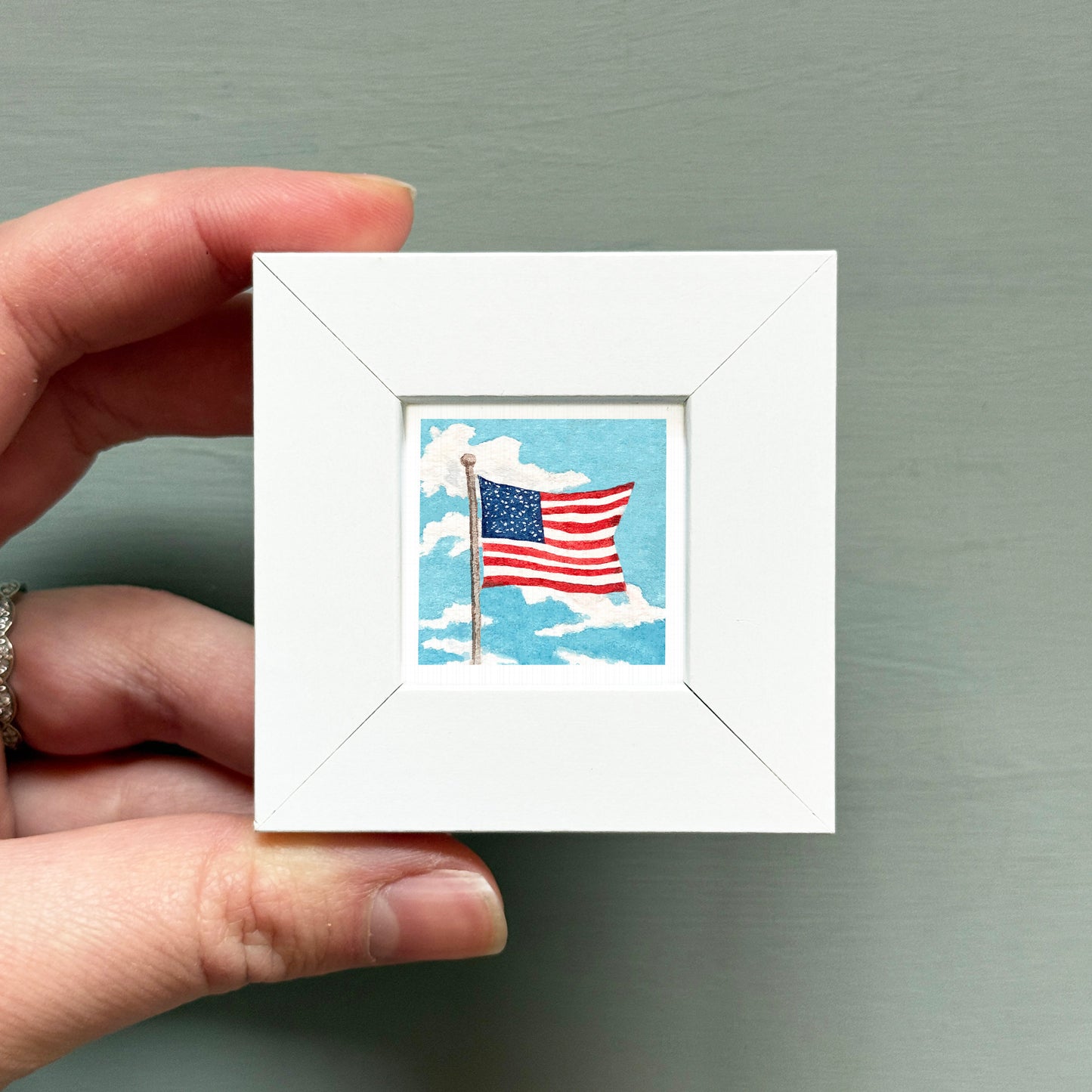 A hand is holding a small white picture frame containing an American flag.